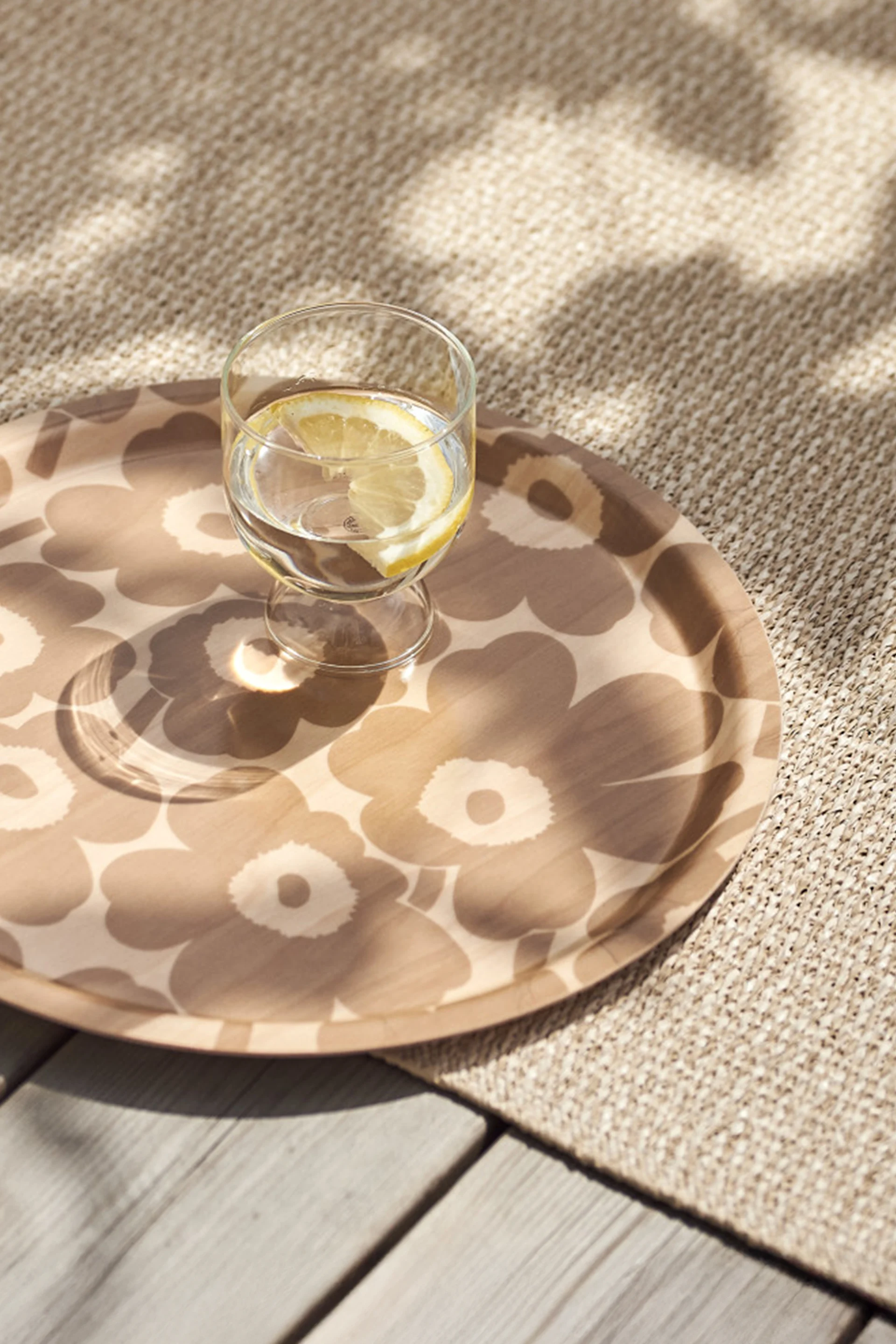Ett glas vatten med citronskiva på en mönstrad rund bricka, placerad på en beige vävd matta och trädäck med fläckigt solljus.