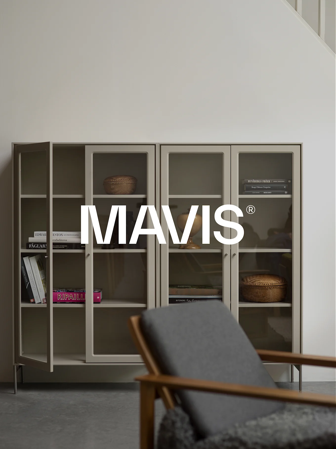 Två ljusgrå skåp med glasdörrar, som visar böcker och inredning. En trästol är suddig i förgrunden. Logotypen 'MAVIS®' är centrerad.