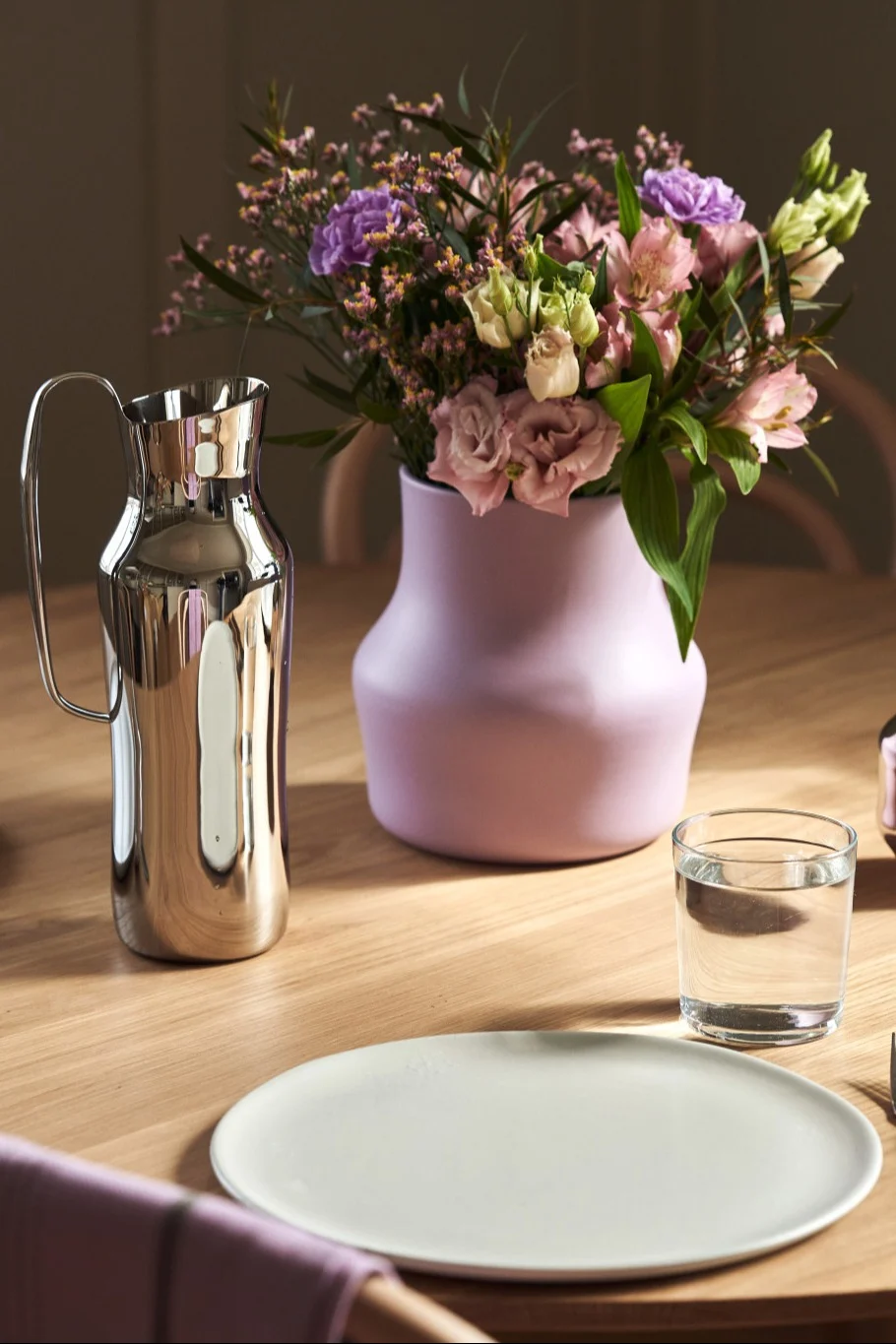 Här ser du den lila Dorotea-vasen med en bukett rosa och lila blommor stå på ett bord bredvid den silverfärgade Dorotea-karaffen.