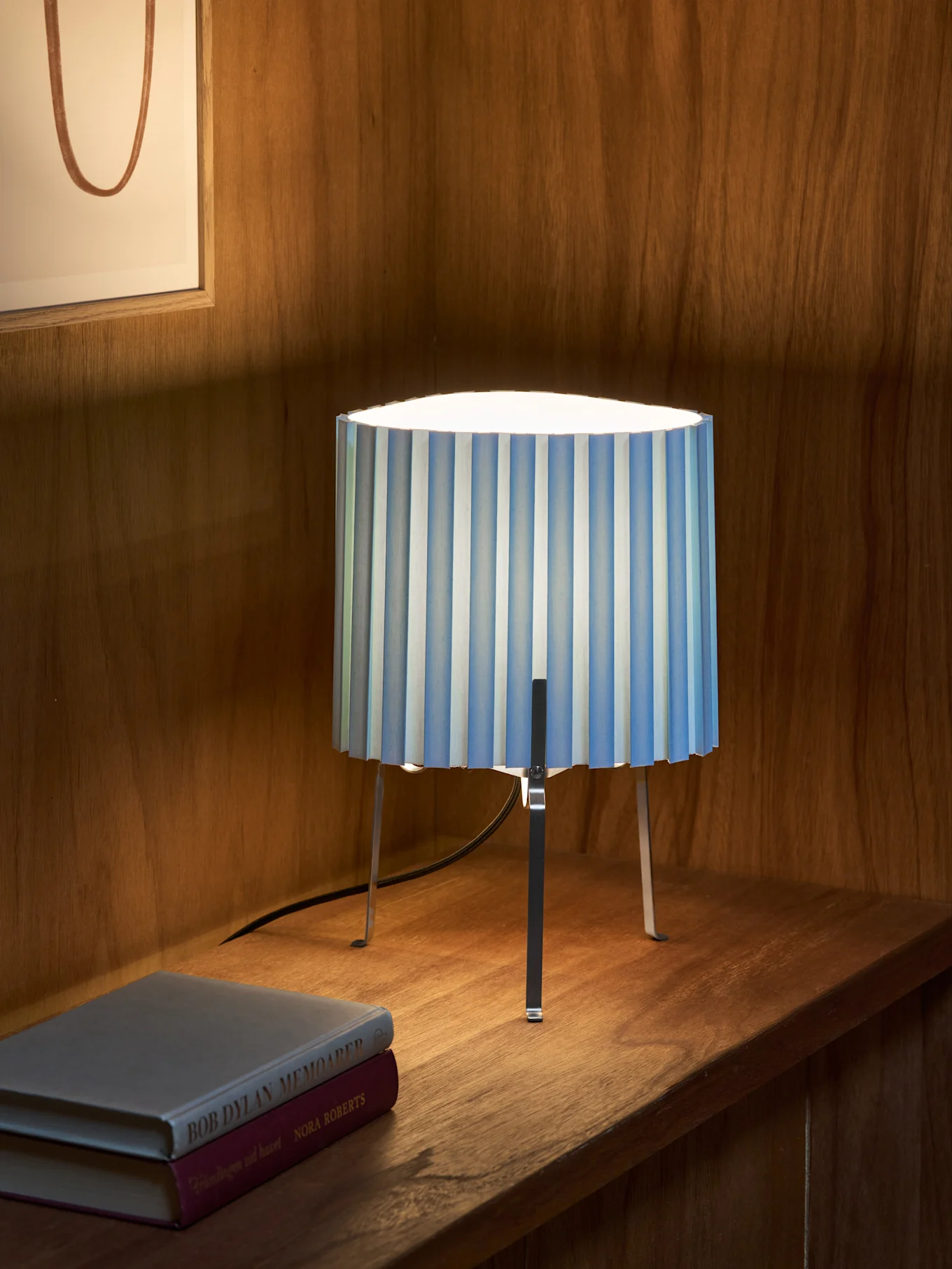 En blå- och vitrandig plisserad bordslampa med tre metallben står på en trähylla bredvid två böcker. Inramad konst i bakgrunden.