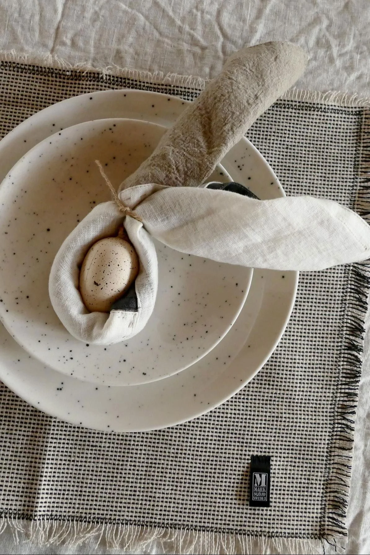 Påskdukning med spräckliga tallrikar, linneservetter vikta som kaninöron med ett spräckligt ägg och en rutig bordstablett.