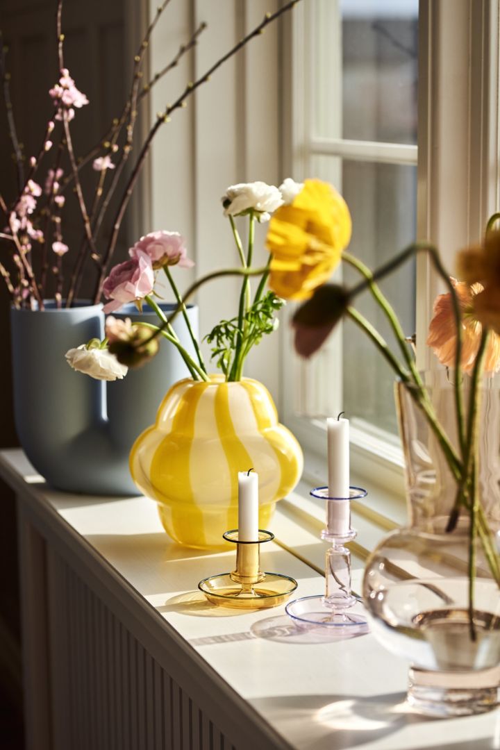 Curlie-vasen från Byon i knallgult står på fönsterbrädan tillsammans med flera andra vaser och ljusstakar i glas. 