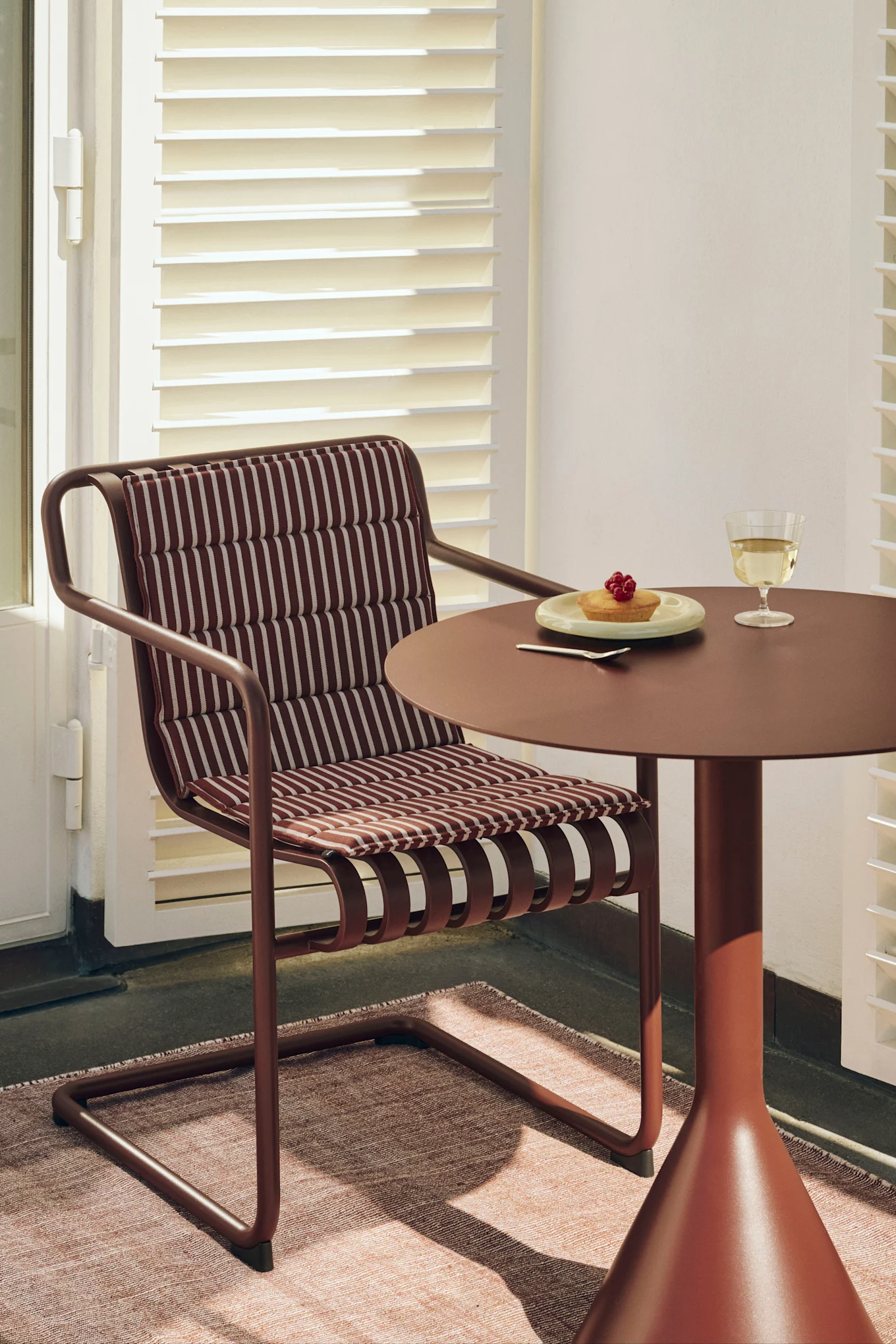 En rödbrun stol med randig dyna intill ett matchande runt bord med en tårta med bär och ett glas vitt vin.