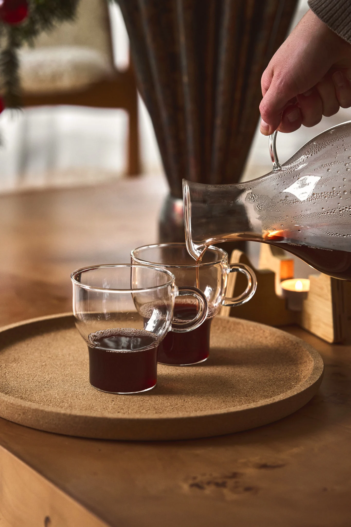 En person häller glögg från en glaskaraff i en glasmugg från Sagaform under ett adventsfika. Allt står på ett träbord med en korkbricka.