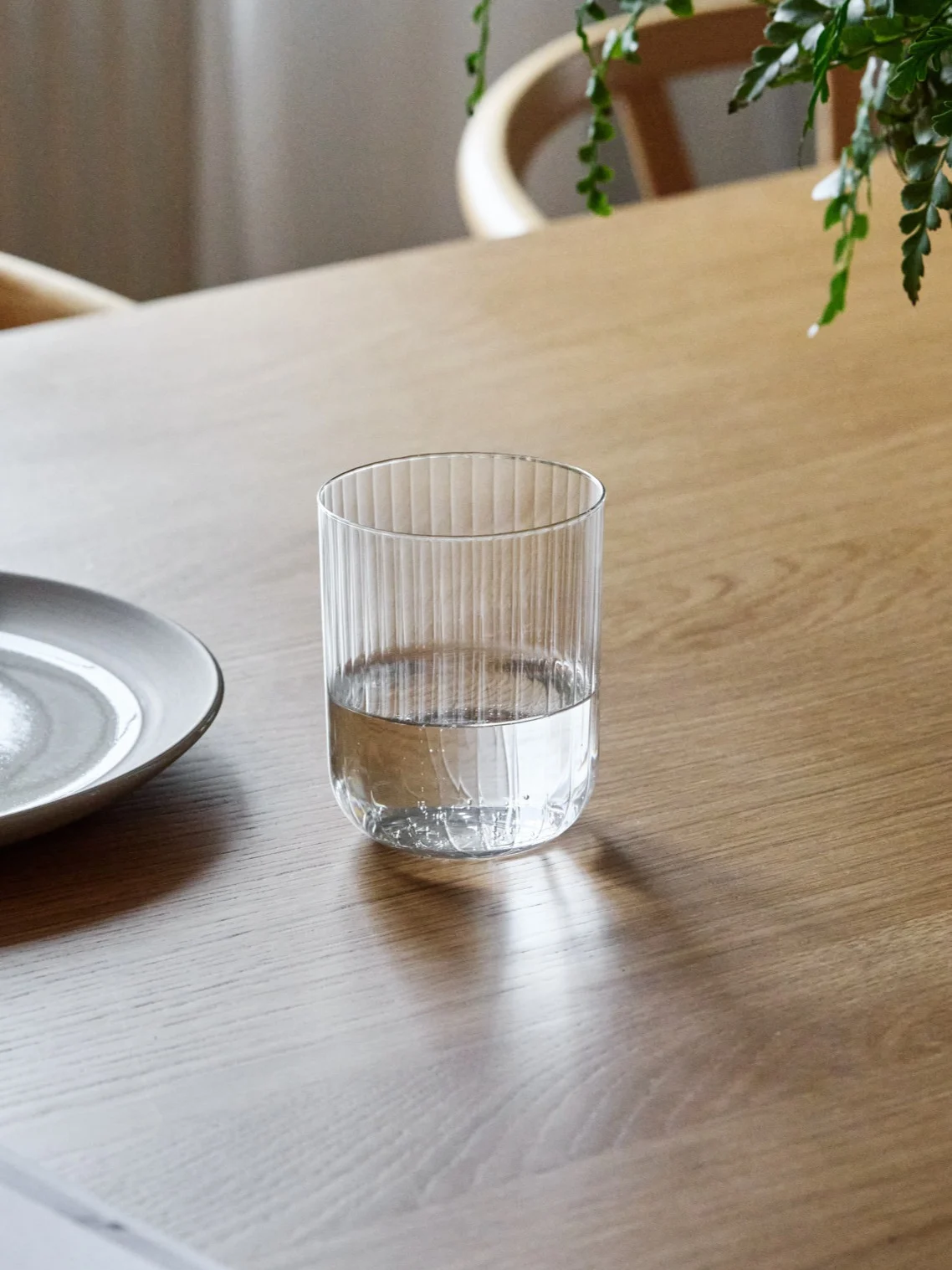 Ett räfflat dricksglas, halvfullt med vatten, står på ett träbord bredvid en keramisk tallrik.
