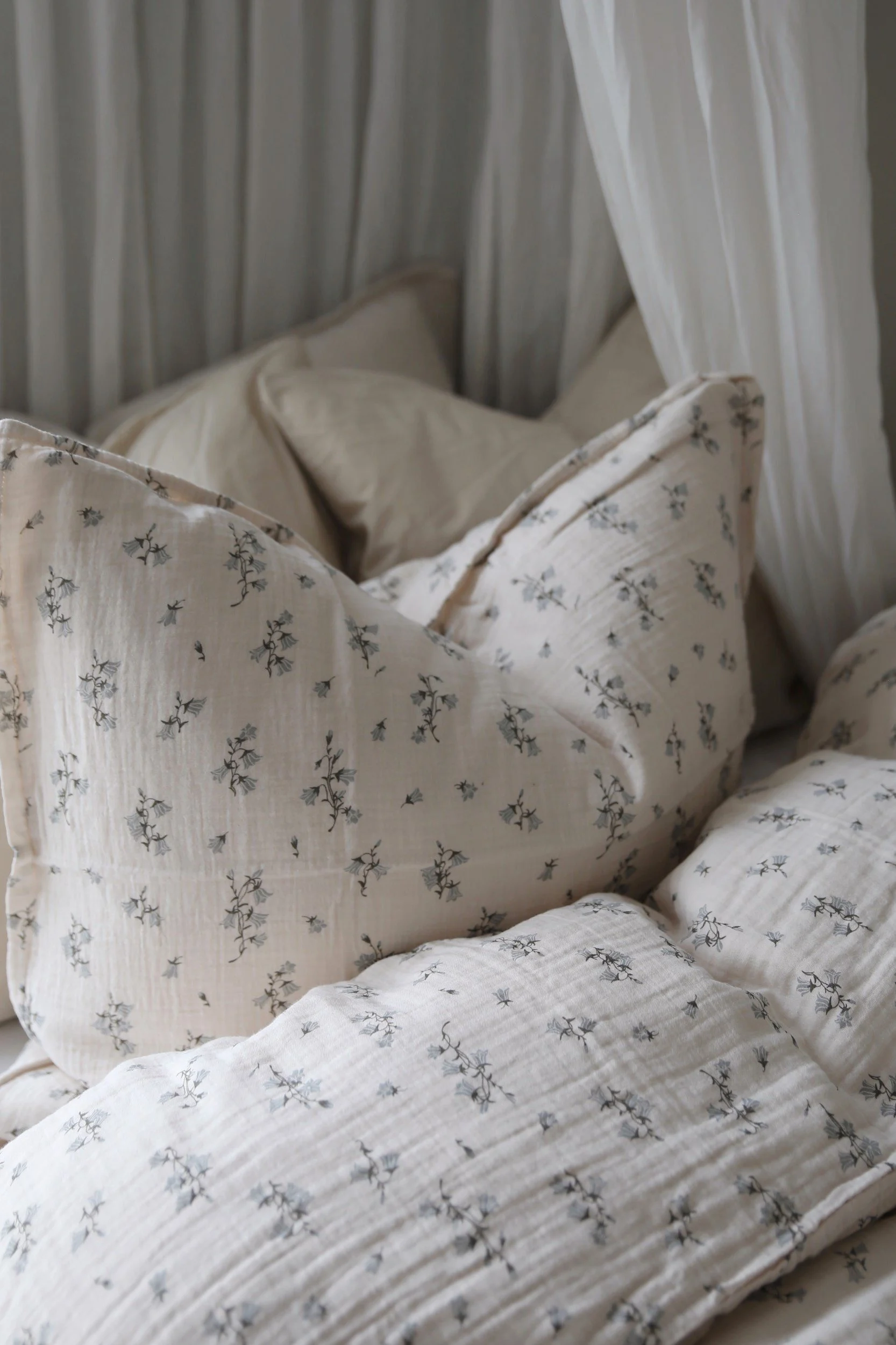Mysiga sängkläder i ljusbeige krinklat tyg med ett delikat blått blommönster och vita gardiner.