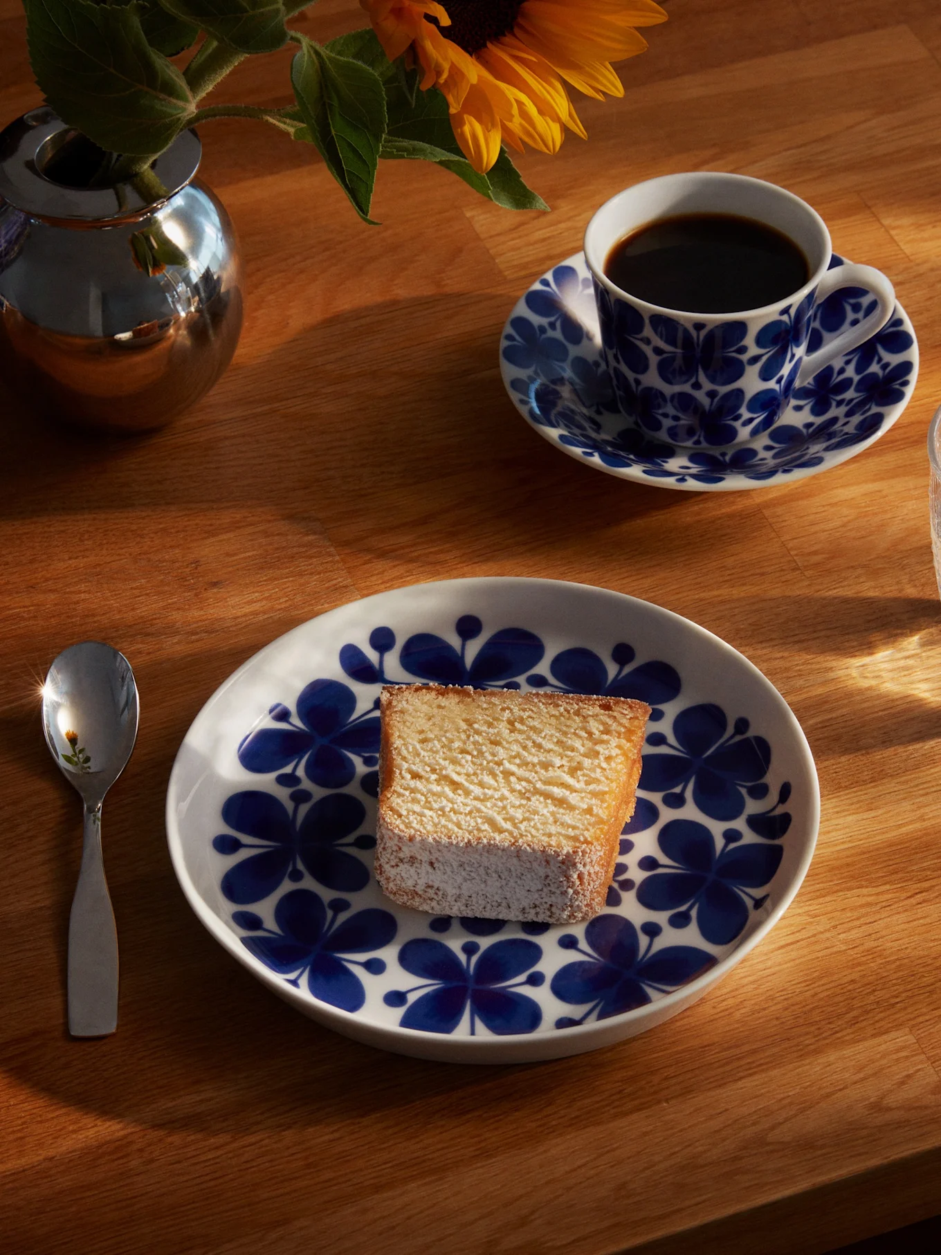 En bit kaka på en blå och vit blommig tallrik, med en kaffekopp, ett glas vatten, en sked och en solros i en silvervas på ett solbelyst träbord.