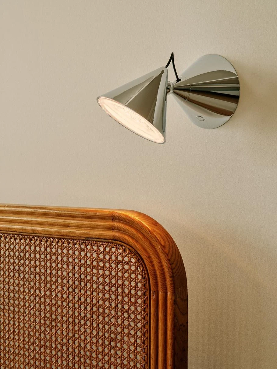Modern kromad vägglampa med konisk skärm som belyser en ljus vägg ovanför en vävd rotting- och trägavel.