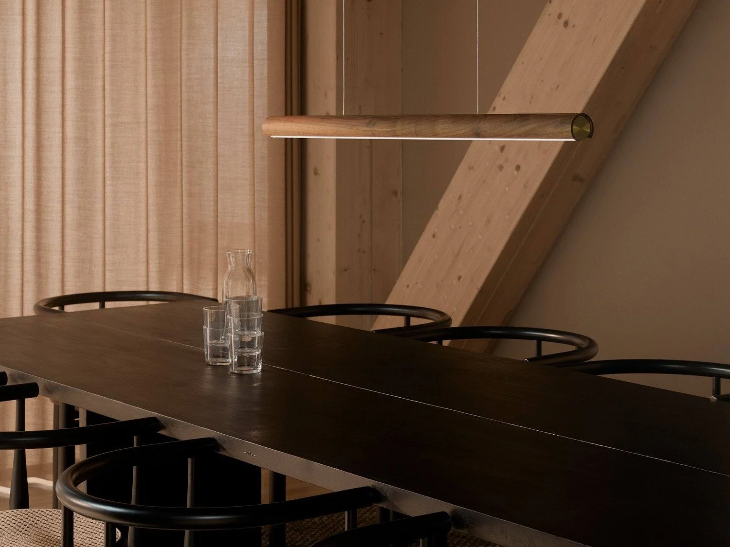 Ett modernt matrum med ett långt mörkt träbord, svarta stolar och en elegant pendellampa i trä. En glaskaraff och tre glas står på bordet.