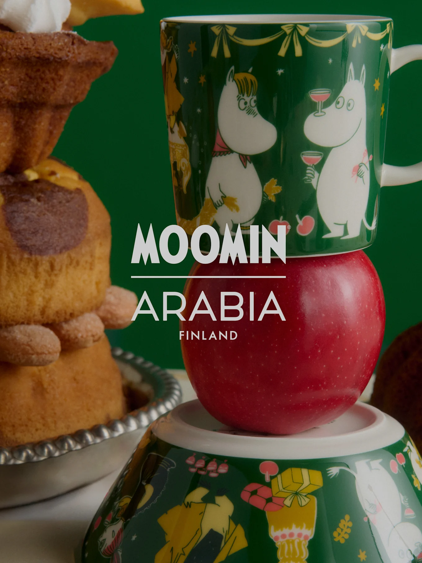 Två staplade ljusblå Muminmuggar med solmotiv på ett träbord med blommor. Text: MOOMIN ARABIA FINLAND.