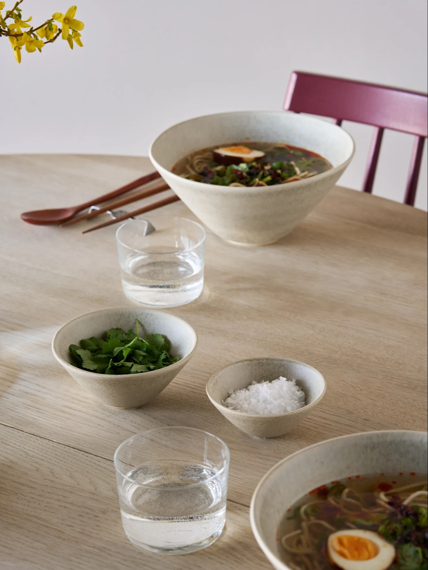 Två skålar ramen med ägg och örter, sidskålar med koriander och salt, vatten, ätpinnar och en sked på ett ljust träbord.