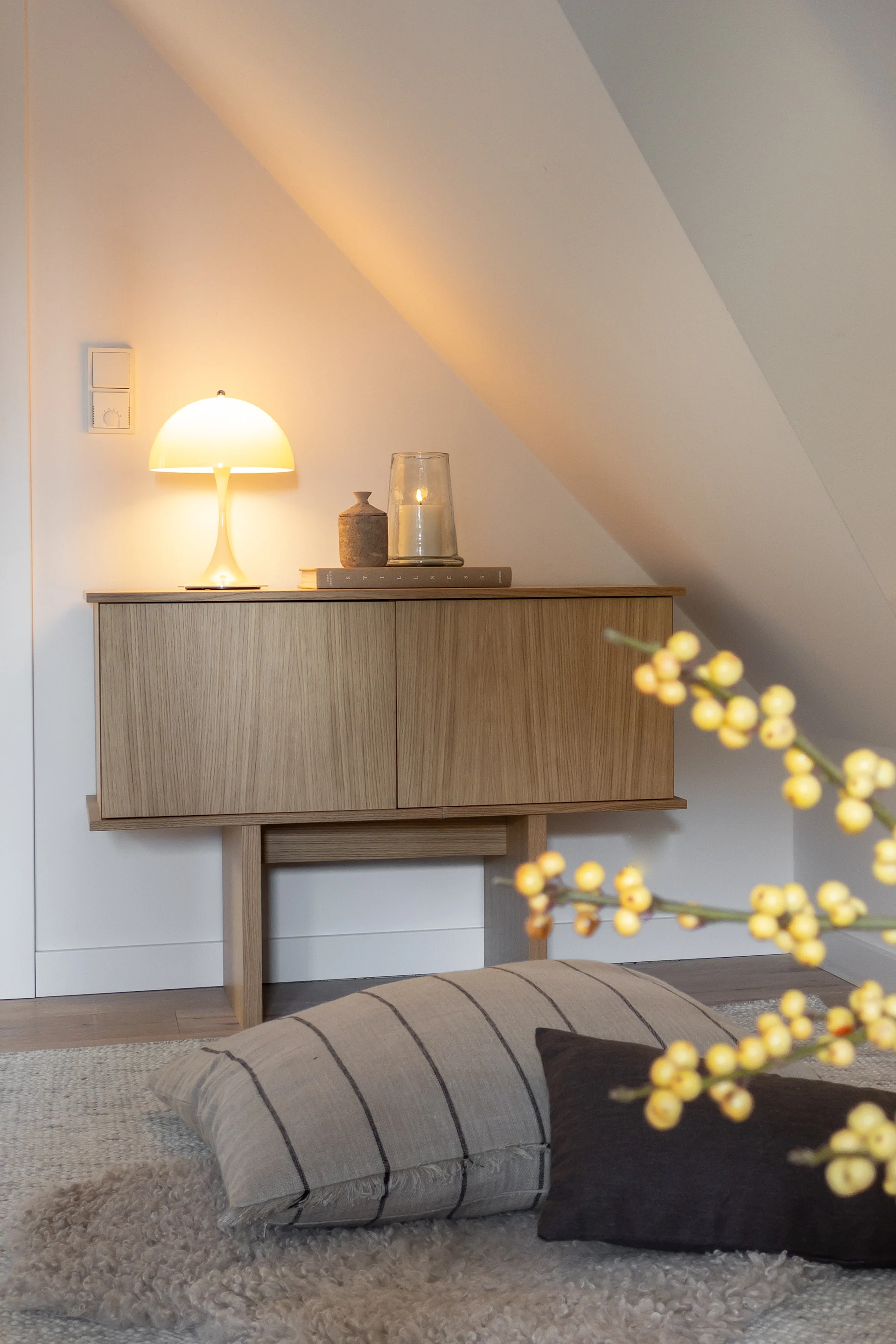 Ett träskåp med en glödande svamplampa och ett tänt ljus, mot en vit, sluttande vägg i ett mysigt rum.