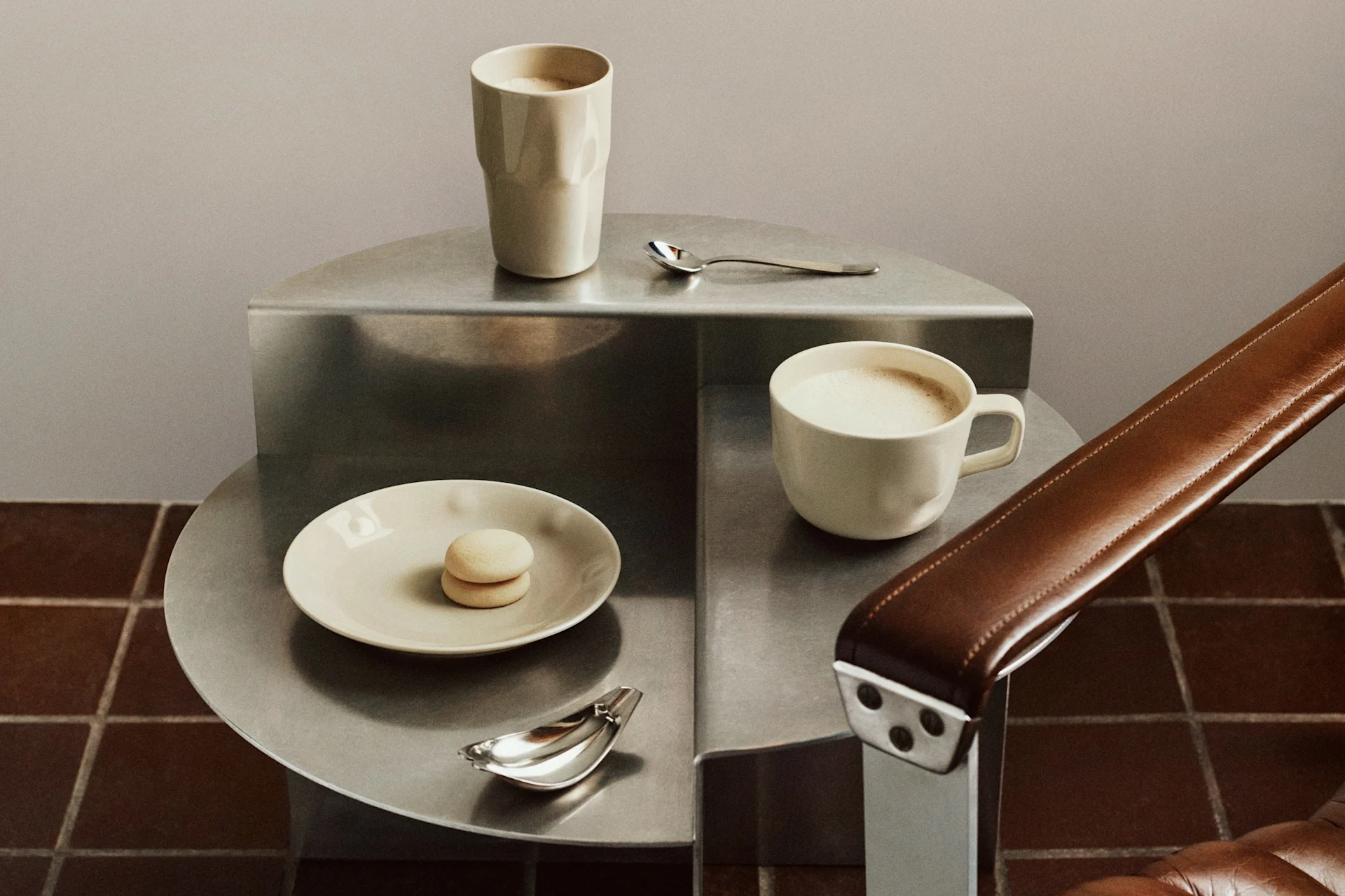 Två ljusa kaffemuggar, en makaron och skedar på ett avsatsbord i metall bredvid en brun läderfåtölj.