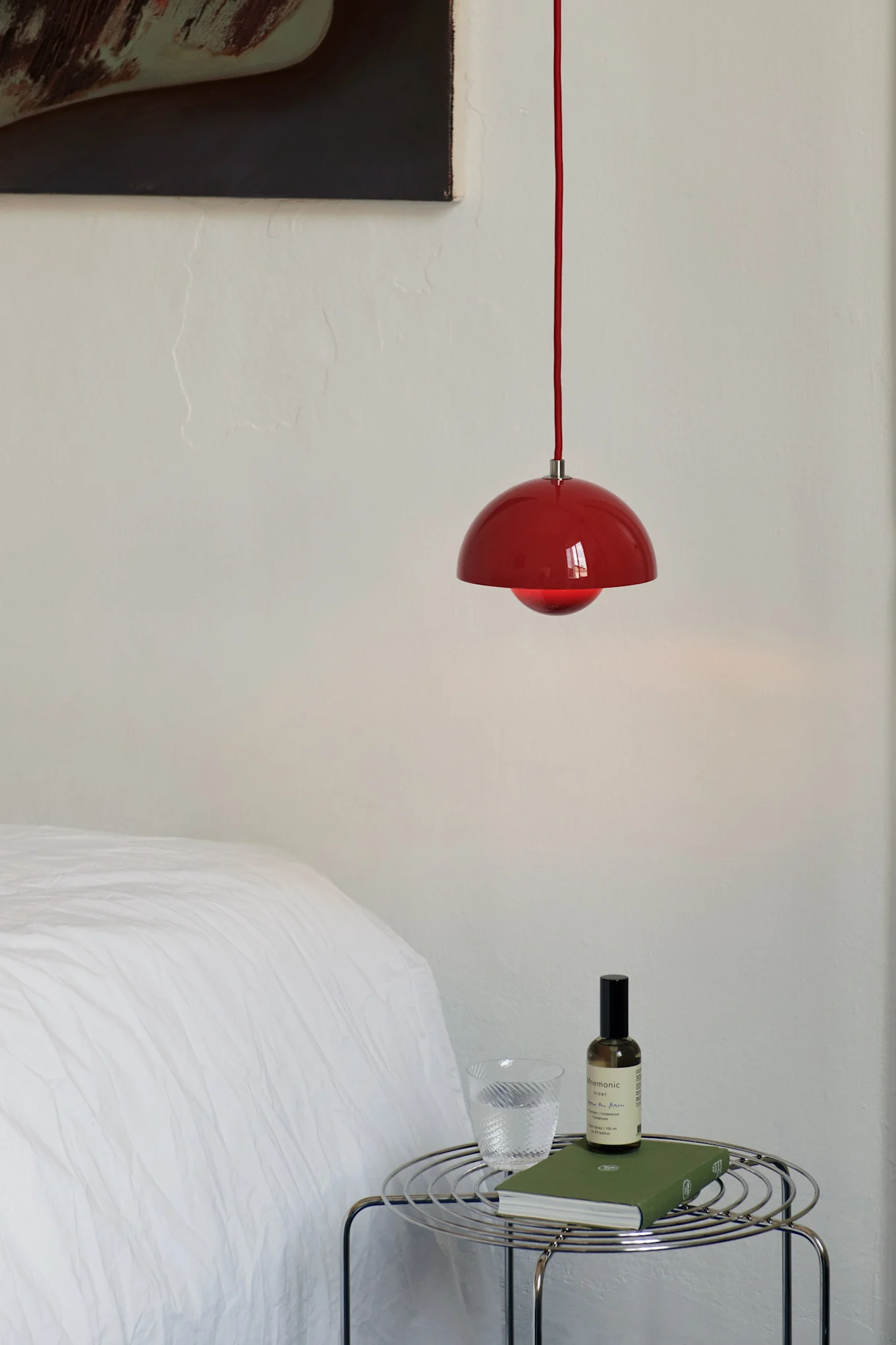 Röd taklampa hänger över ett kromat sidobord i tråd med ett glas vatten, en grön bok och en sprayflaska i ett sovrum.