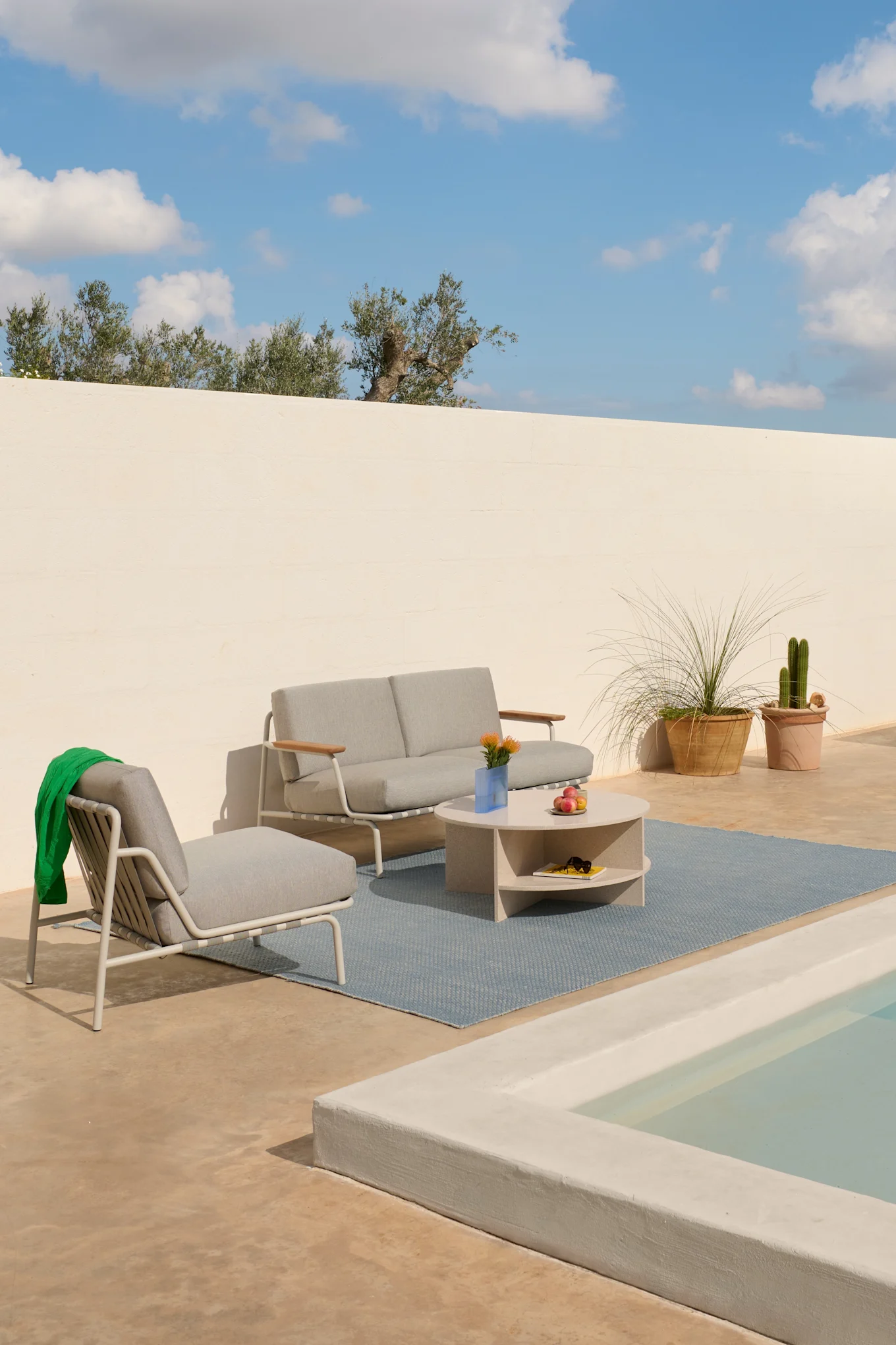 Utomhusloungeset med ljusgrå soffa, fåtölj och runt soffbord på en blå matta, bredvid en liten pool.