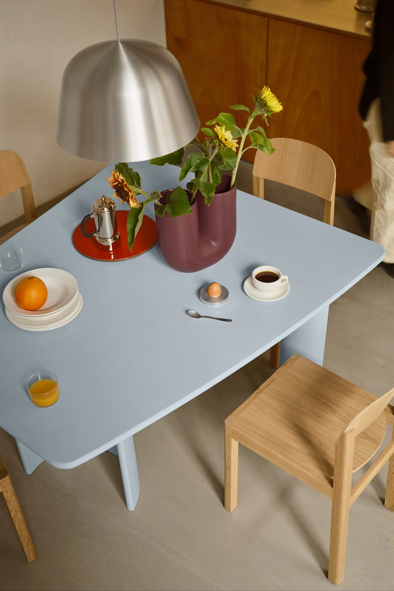 Ett blått matbord dukat för frukost med kaffe, apelsinjuice, ett ägg och solrosor i en vinröd vas, under en silverlampa.