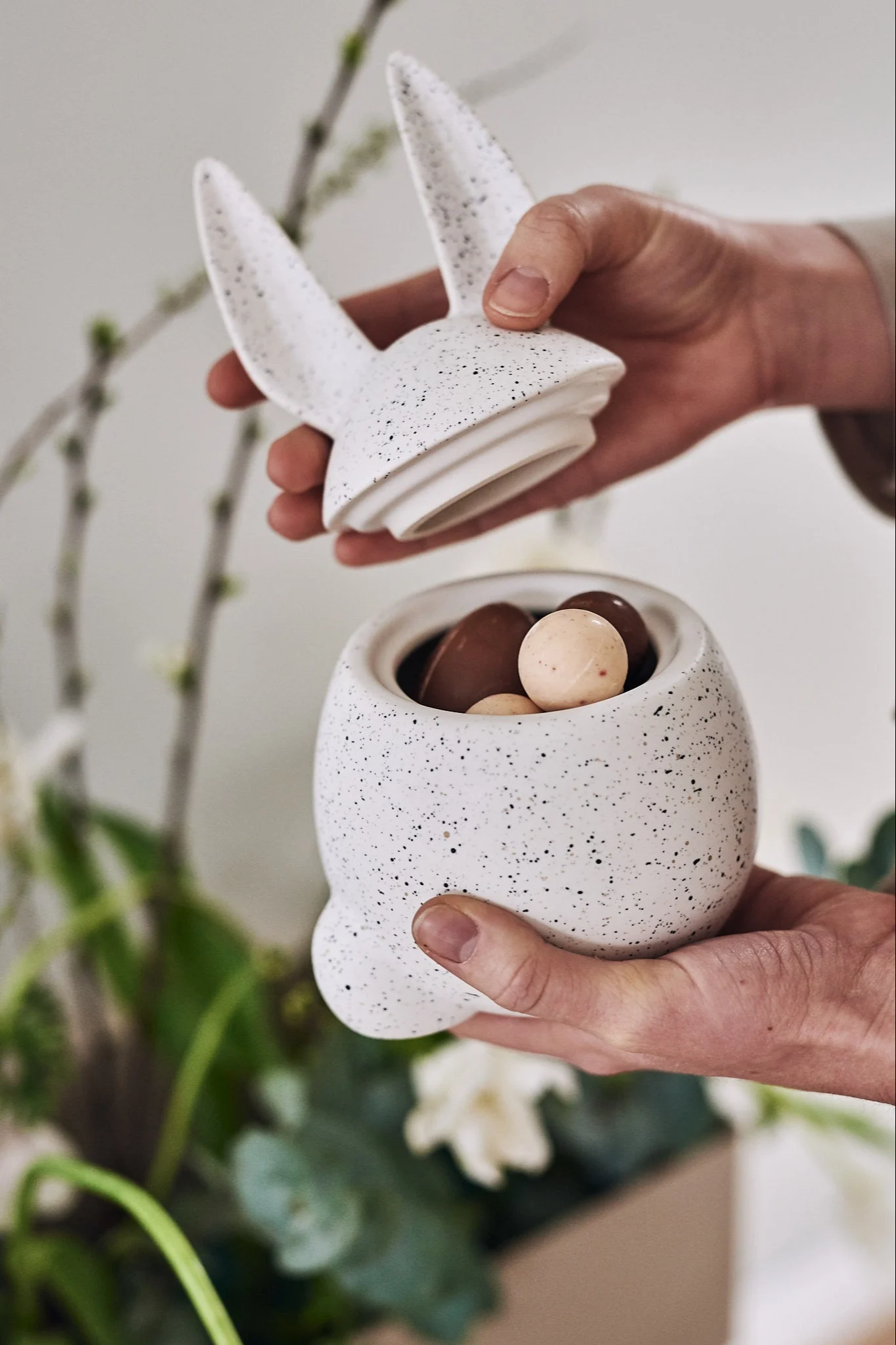 Händer öppnar en vit, fläckig kaninbehållare fylld med chokladpåskägg.