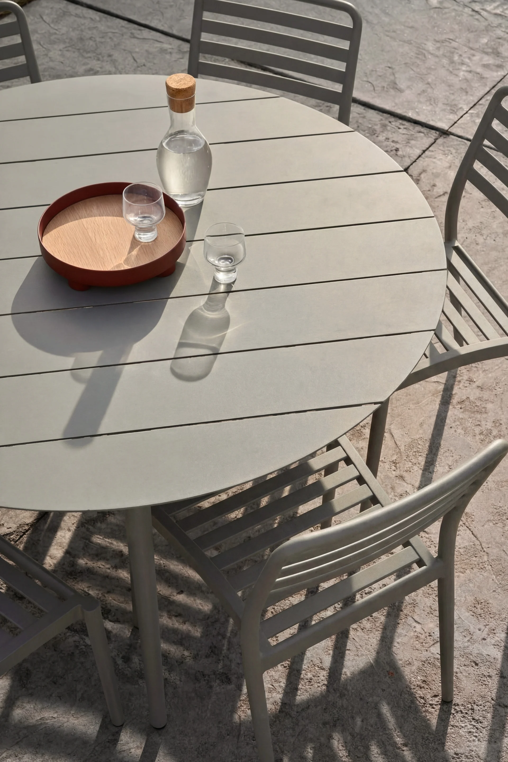 Runt grått utomhusbord, tre stolar. Glaskaraff vatten och två glas, det ena på träbricka, står på bordet.