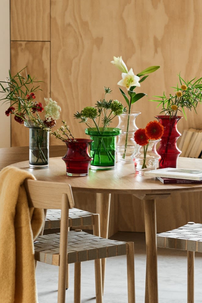 En samling Kosta Boda-vaser i klart, rött och grönt glas står på ett matsalsbord med färska snittblommor. 