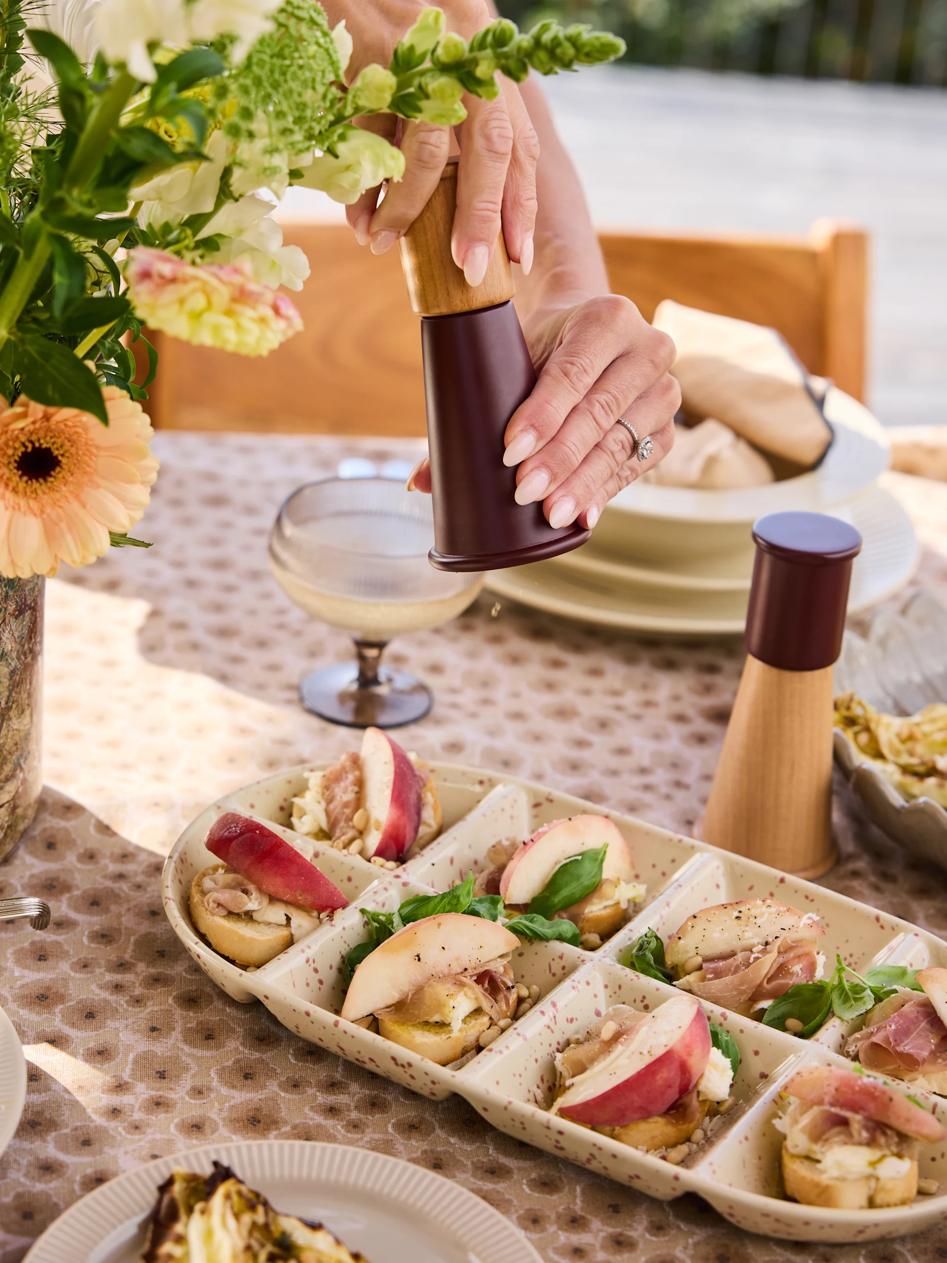 Händer maler peppar på crostini-fat med persikor, prosciutto, ost, pinjenötter, basilika.