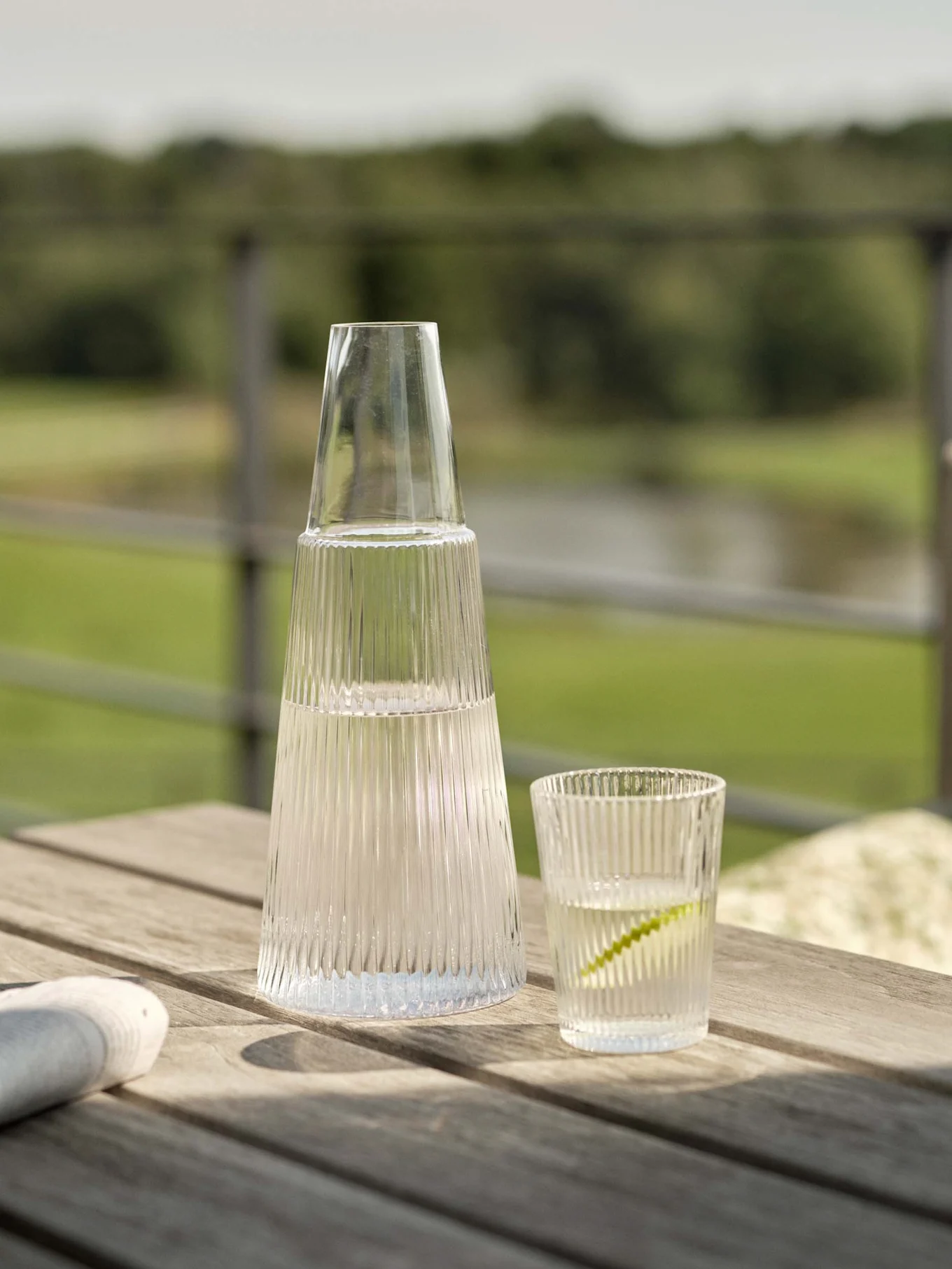 Klar räfflad glaskaraff och matchande glas med vatten och lime på ett utomhusbord i trä.