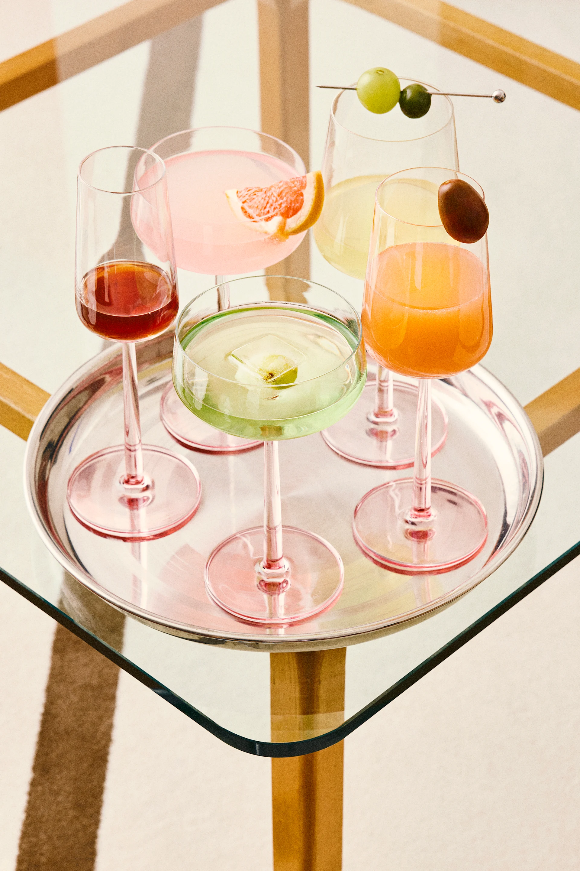 Fem färgglada cocktails på en silverbricka, inklusive rosa, gröna, orange och röda drinkar med olika garnityr, på ett glasbord.