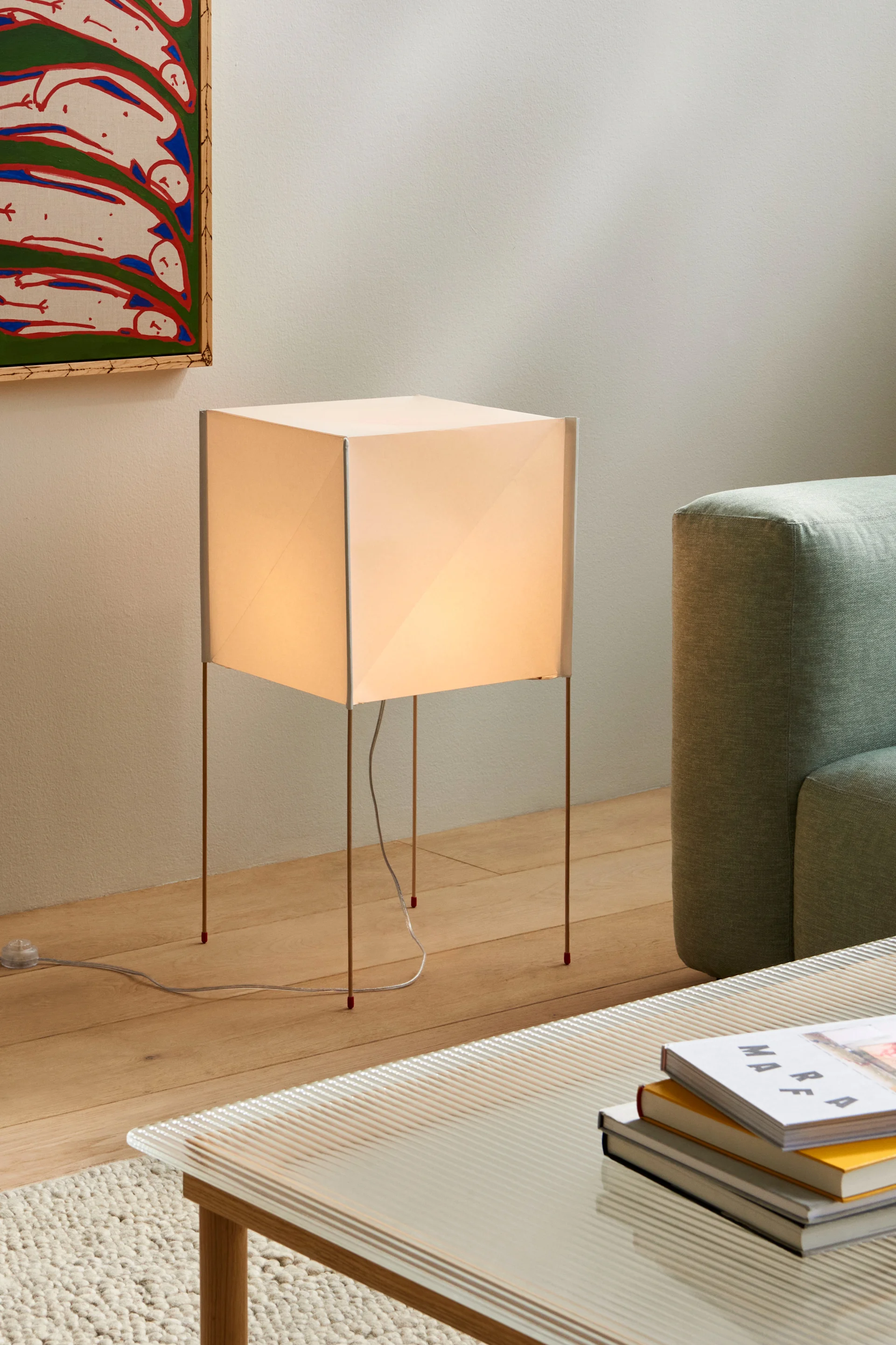 En glödande kvadratisk golvlampa med tunna ben intill en ljusgrön soffa och ett soffbord med böcker.