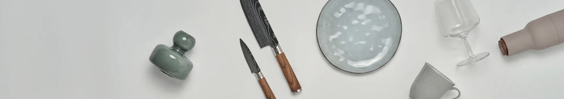 Olika köks- och matvaror, inklusive en grön vas, två knivar, en fläckig tallrik, ett vinglas, en beige flaska och en mönstrad mugg.