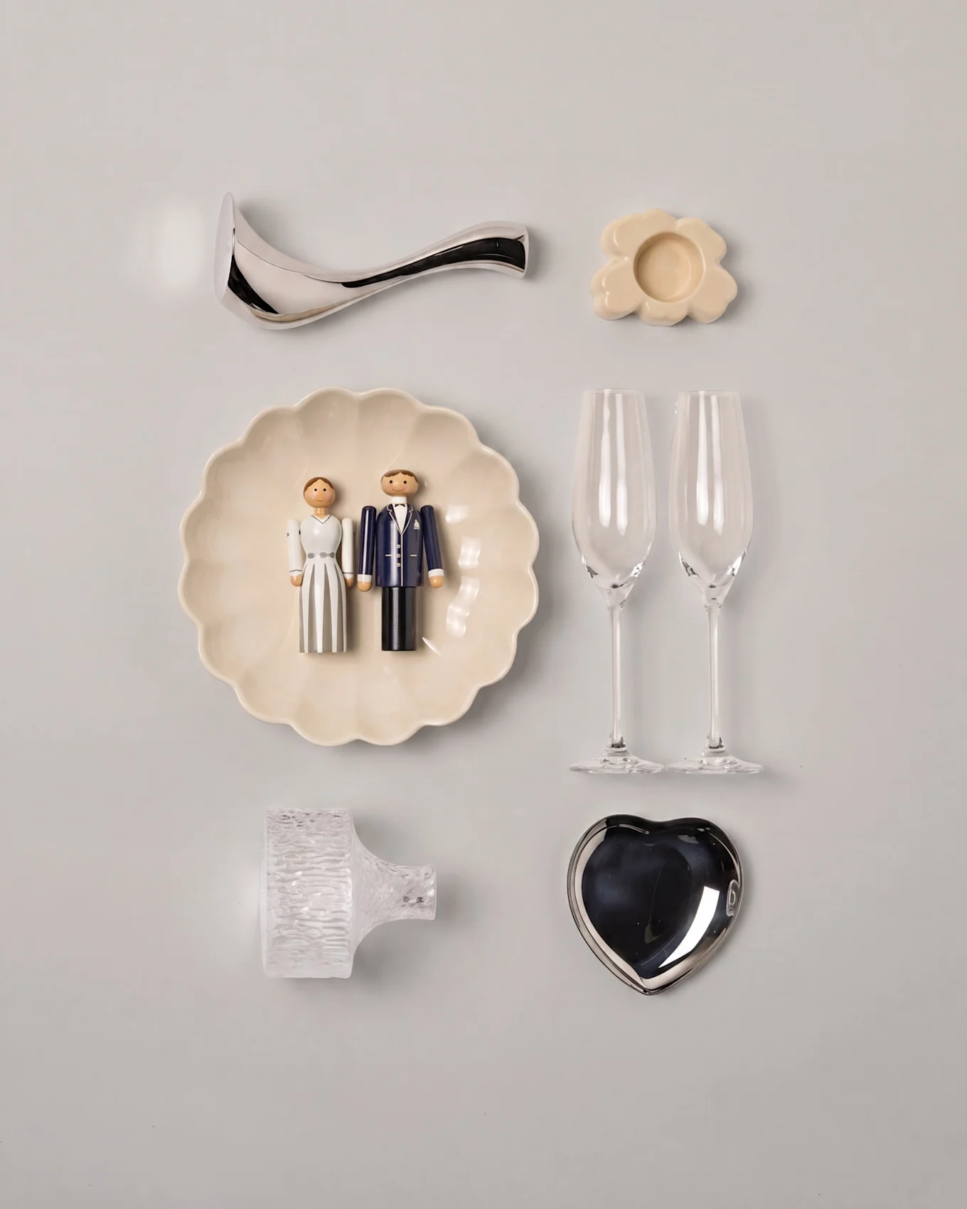 Bröllops-flat lay med träfigurer av brudpar på en räfflad tallrik, två champagneglas, ett silverfärgat hjärtfat och diverse dekorationer.