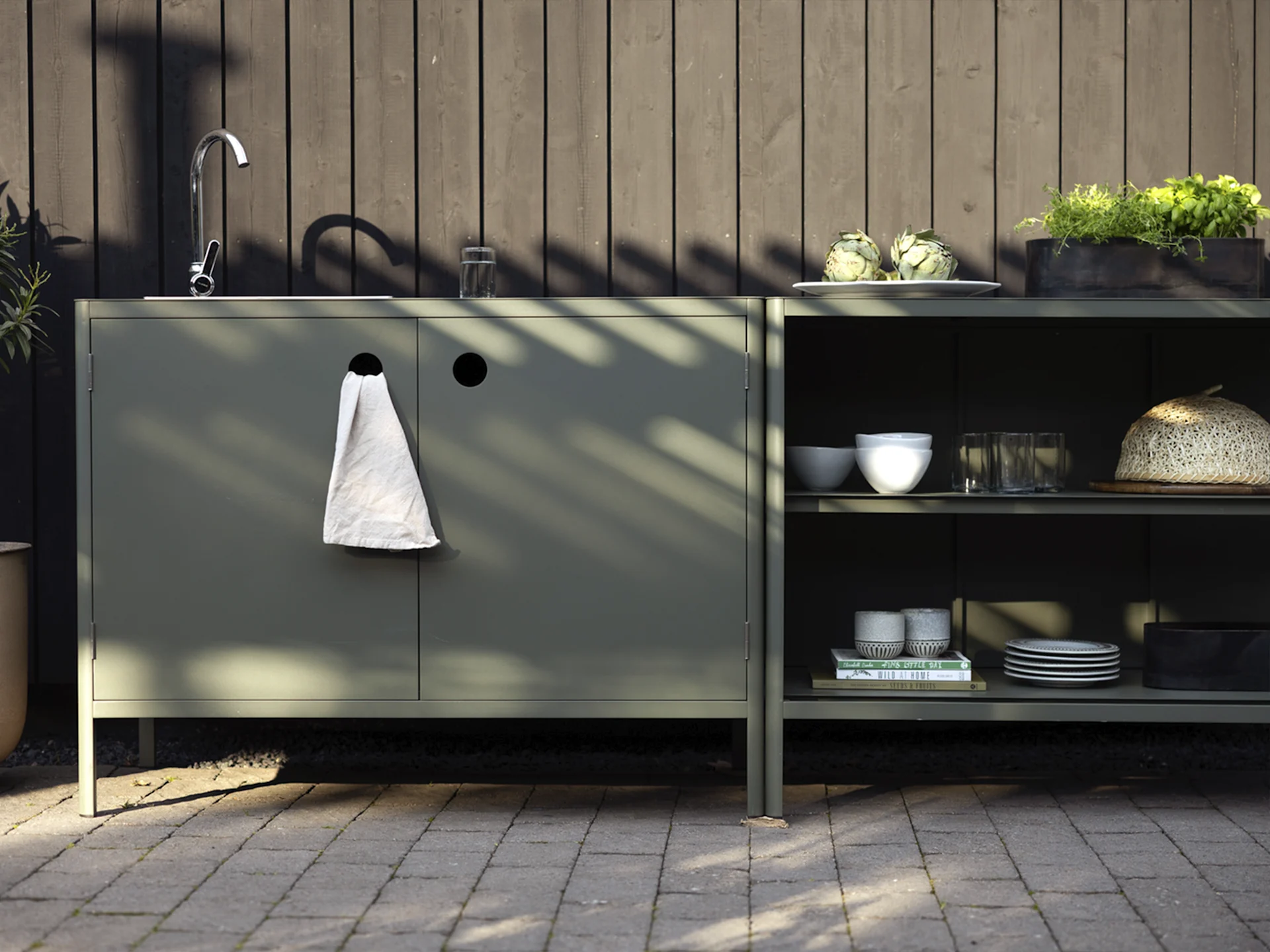 Utomhuskök med mörkgröna skåp, diskho, kran och hyllor med kronärtskockor, örter och porslin.