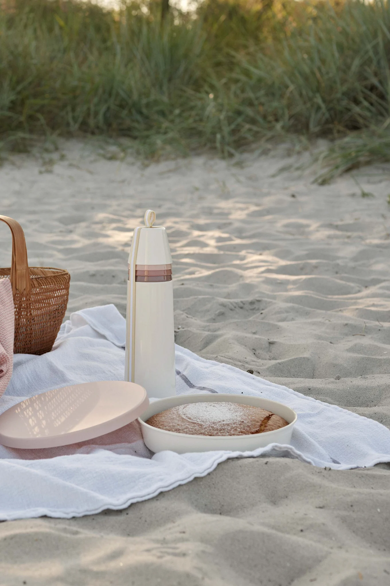Picknick på sandstrand med krämfärgad termos, kaka med florsocker och en flätad korg.