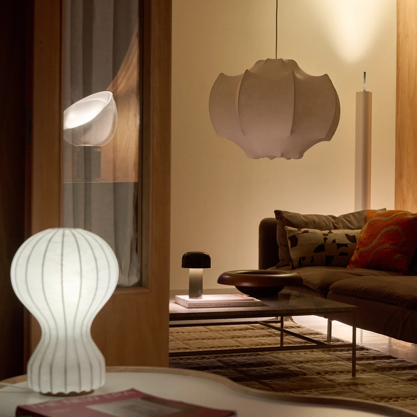 Ett vardagsrum med en brun soffa och soffbord, upplyst av en vit räfflad bordslampa, en mörk svamplampa och en stor veckad taklampa, som skapar ett varmt sken.
