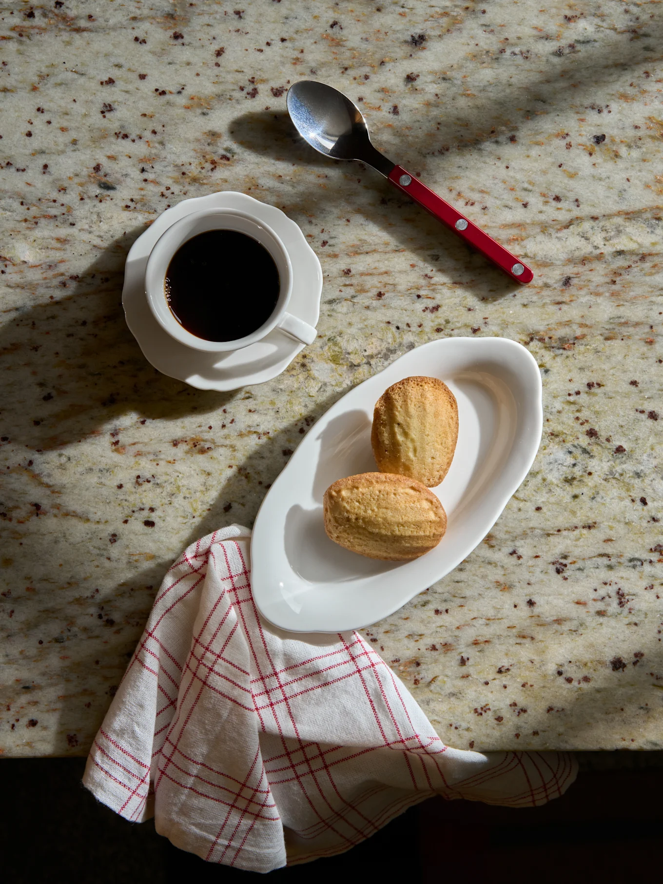 Övervy: svart kaffe i vit kopp, två madeleinekakor på vit tallrik, sked och randig servett på marmorbänk.