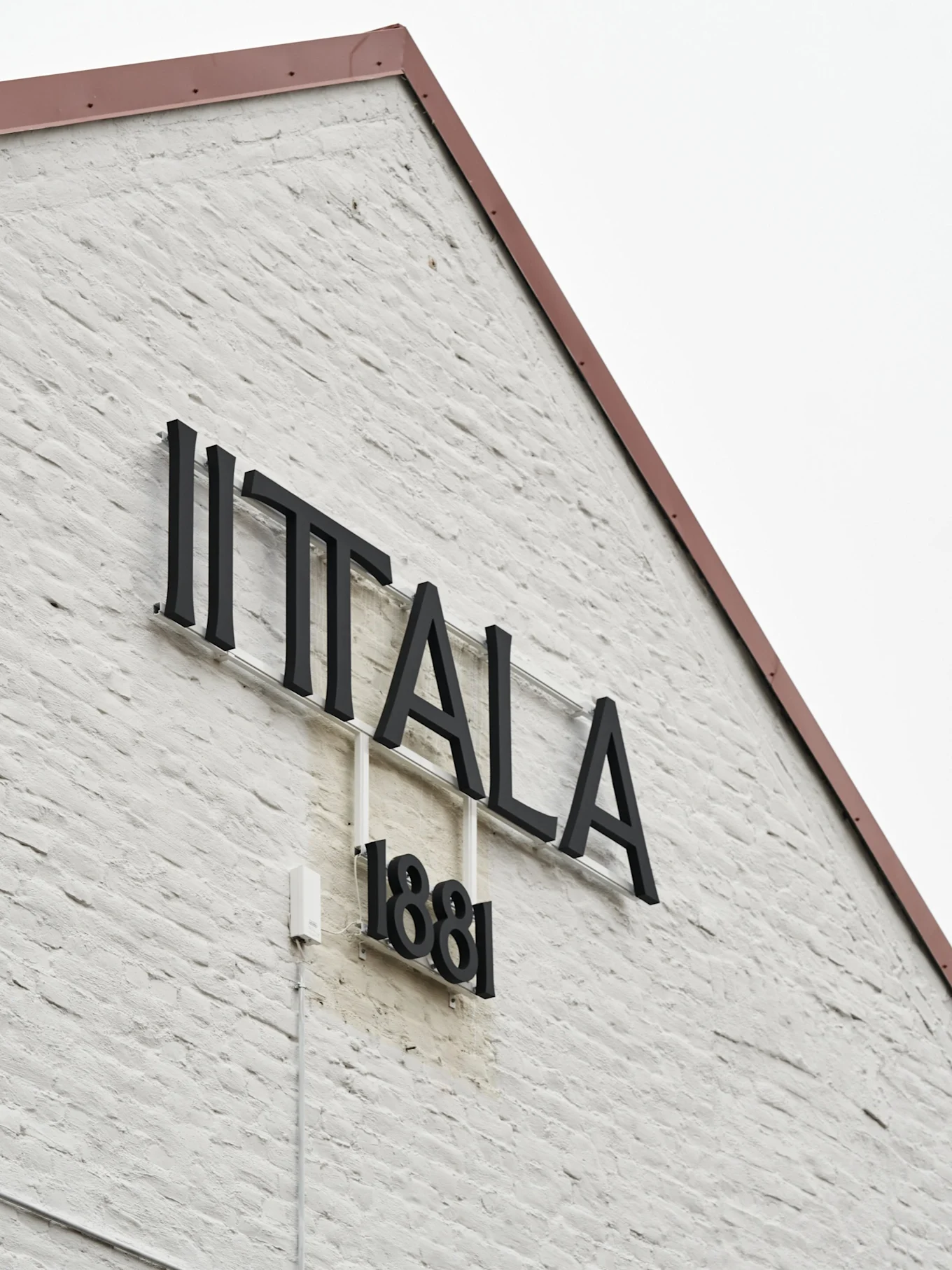 Iittala-logotypen och '1881' på en vit tegelvägg med ett rödbrunt tak.