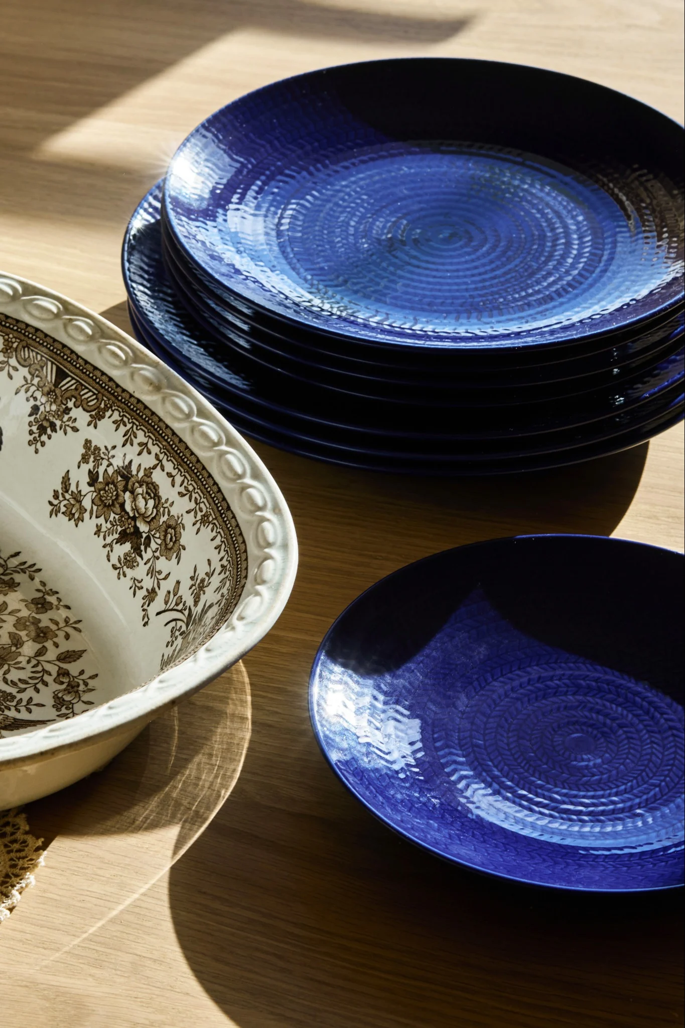 Stapel djupt blå, texturerade tallrikar från Rörstrand Blå Eld, bredvid ett utsmyckat vitt serveringsfat med brunt blommönster på ett träbord.