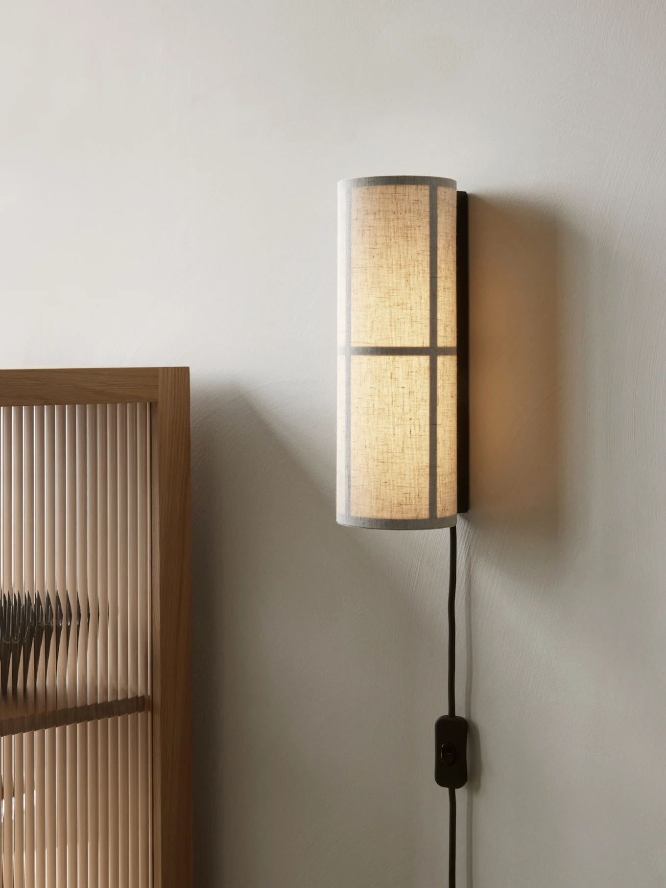Cylindrisk vägglampa med en genomskinlig tygskärm, monterad på en ljus vägg intill en träribbskärm.