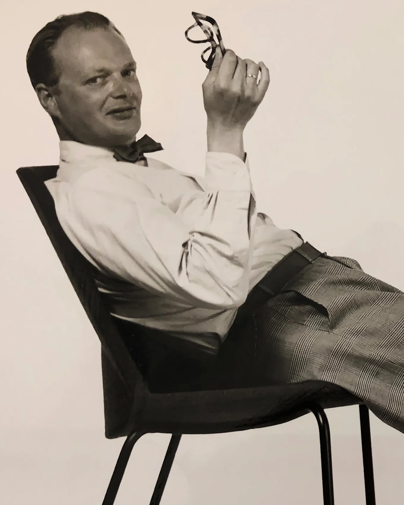 Man i fluga och rutiga byxor lutar sig tillbaka i en stol, håller glasögon och ler mot kameran.