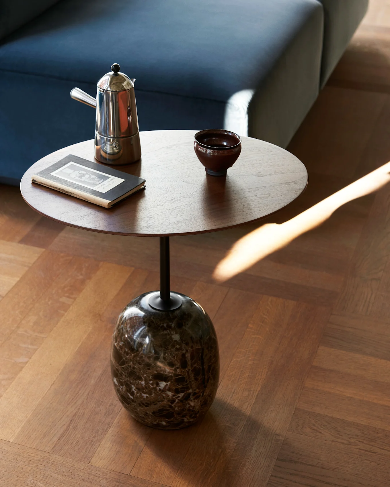 Runt sidobord i trä med marmorfot, med en mokkabryggare, mörk skål och gammal bok, bredvid en blå soffa.