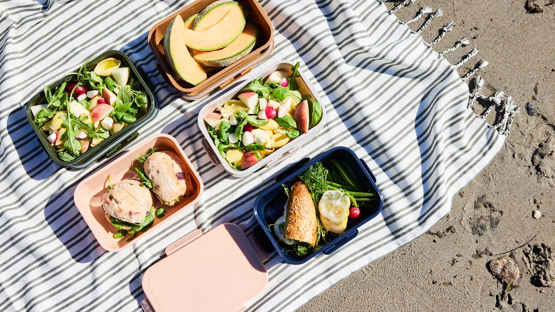 Picknickutbredning på en randig filt på sanden, med flera lunchlådor innehållande frukt, pastasallad och smörgåsar.