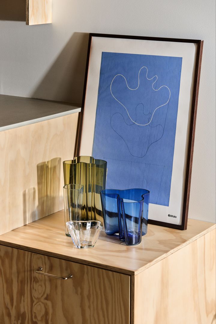 Iittalas ikoniska vaser designade av Alvar Aalto i olika färger står på ett sidobord i trä framför en tavla med blått tryck. 