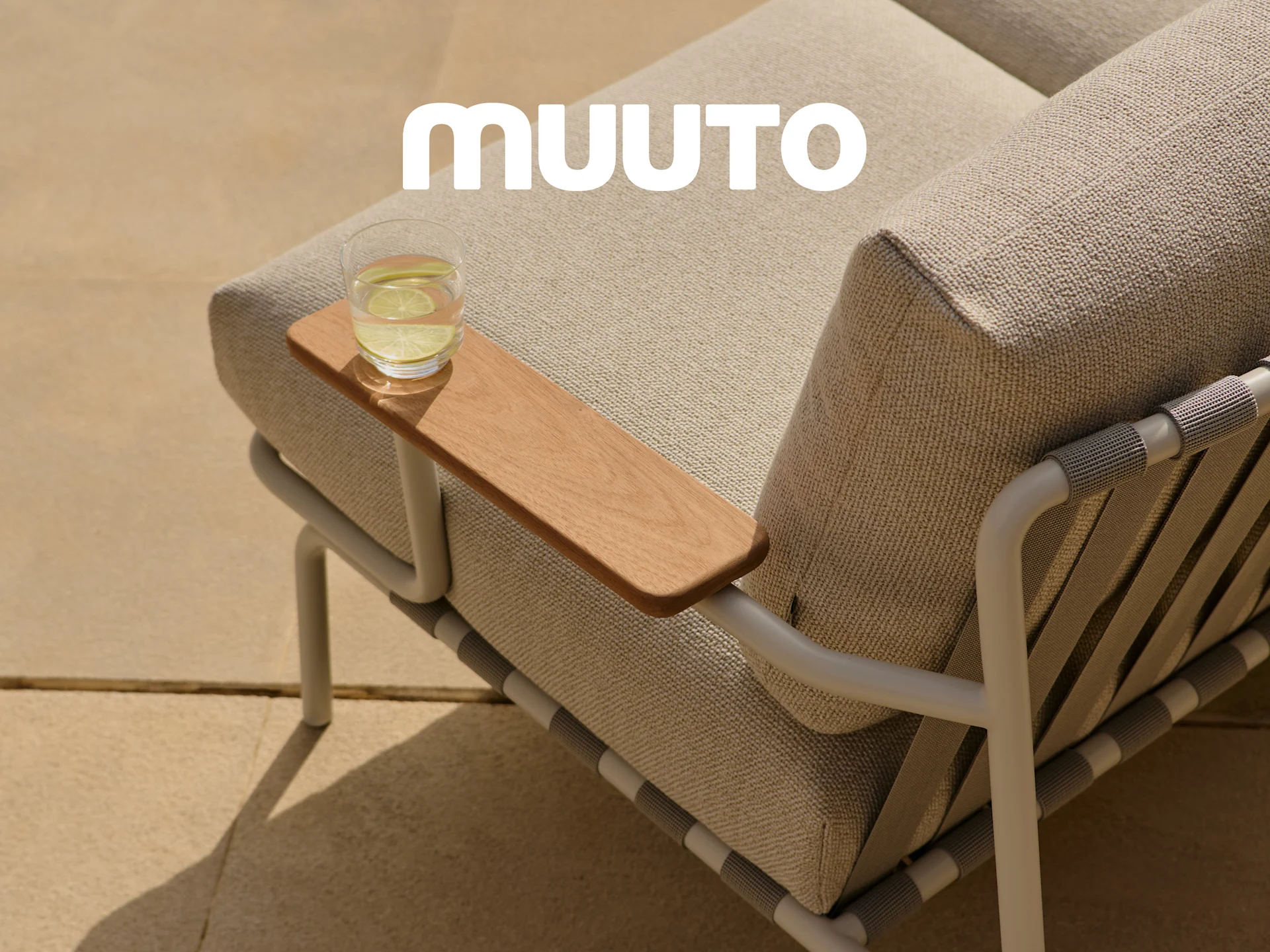 Närbild av en utomhussoffa med ljusa tygkuddar, ett träarmstöd med ett glas vatten och den vita Muuto-logotypen.