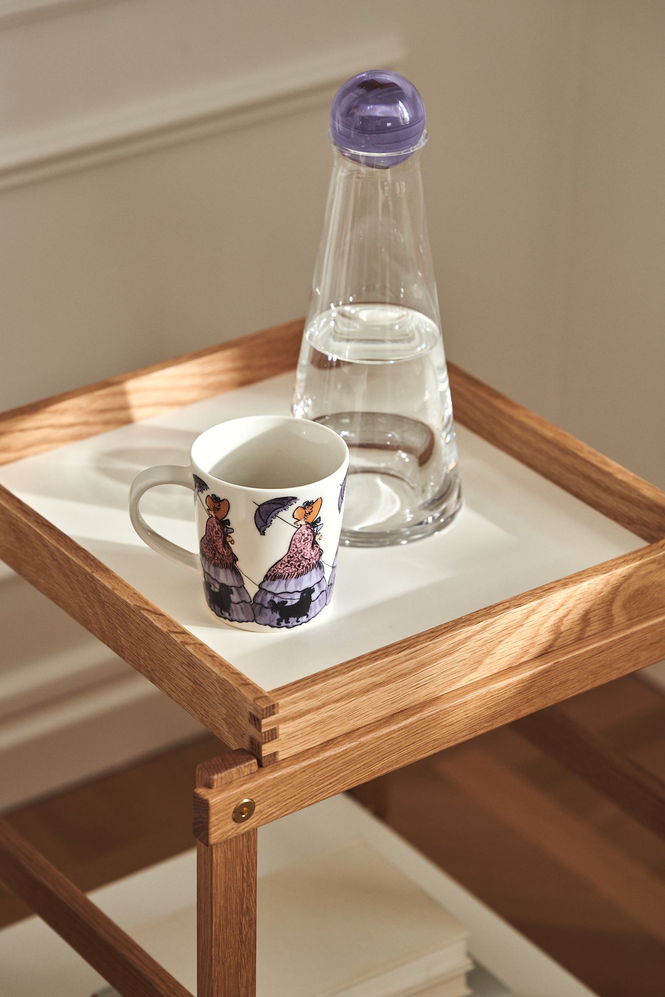 Mugg med Elsa Beskow-motivet Tant Gredelin från Design House Stockholm samt Fia-karaff i glas med lila lock på Frame-hylla i ek och vitt. 