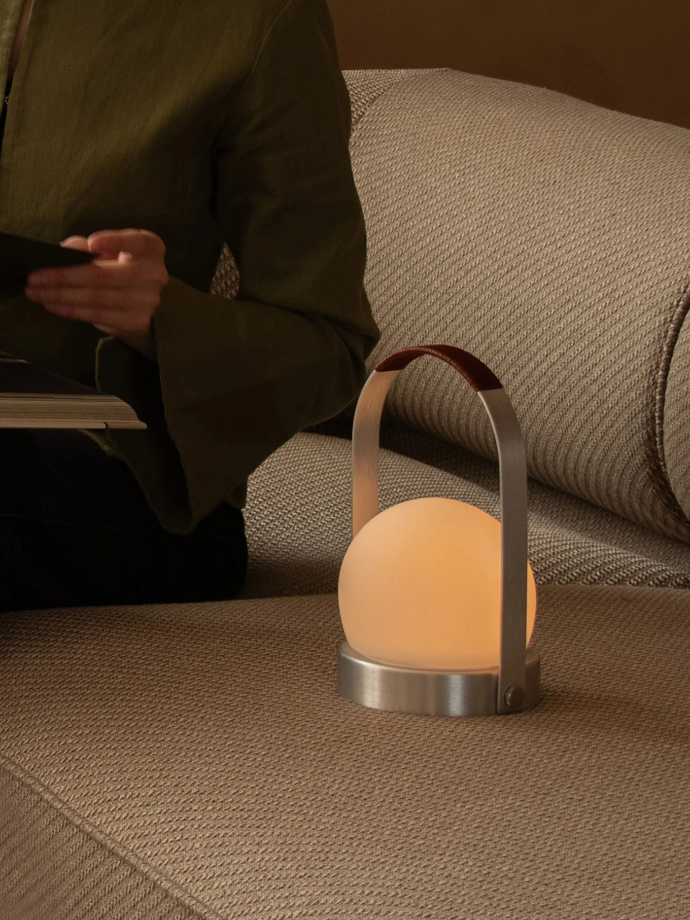 Person läser på soffa med en glödande portabel lampa med frostad glob, metallfot och läderhandtag.