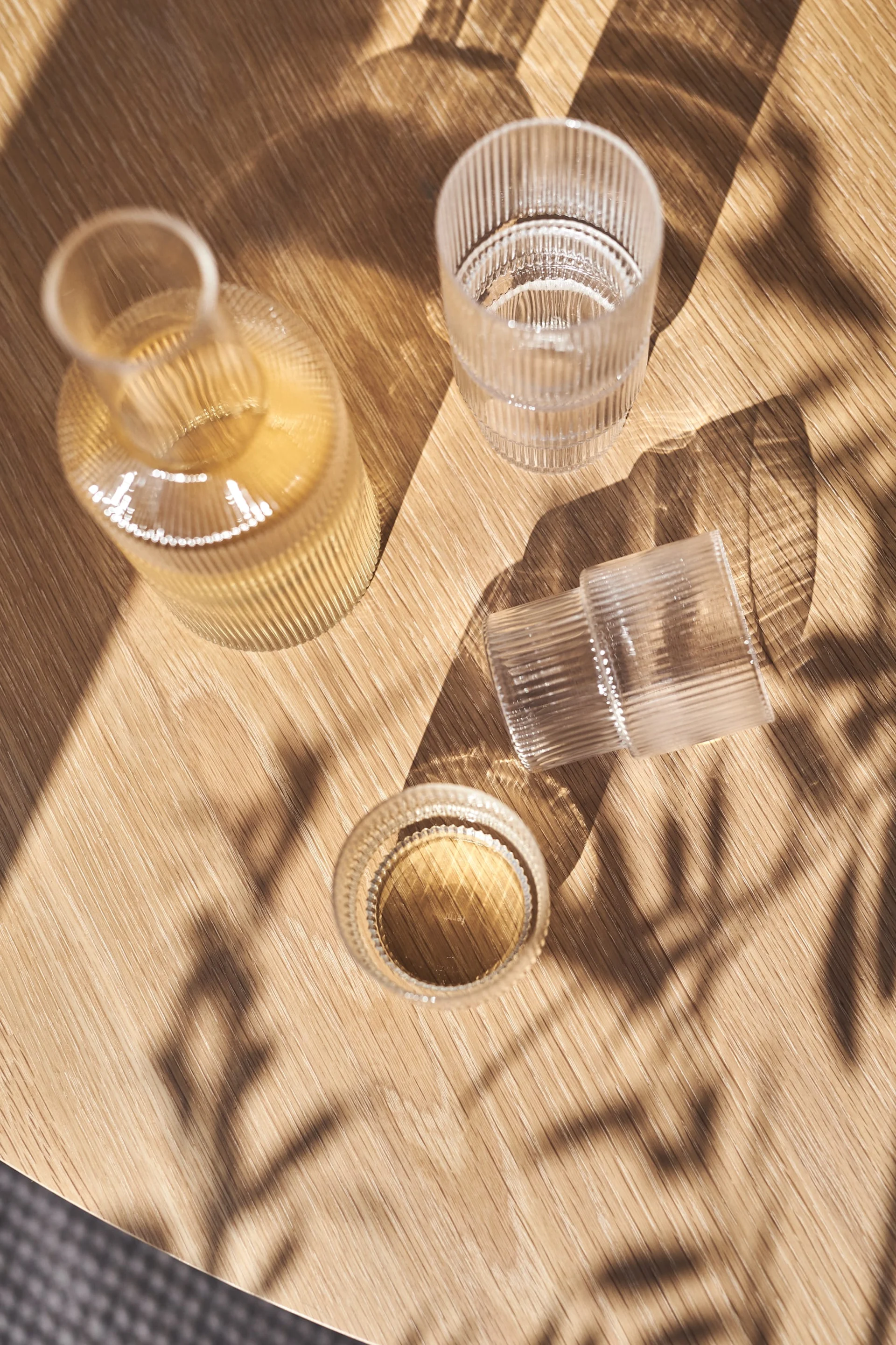 En räfflad glasflaska och två räfflade glas, ett med vätska, på ett solbelyst träbord med bladskuggor.