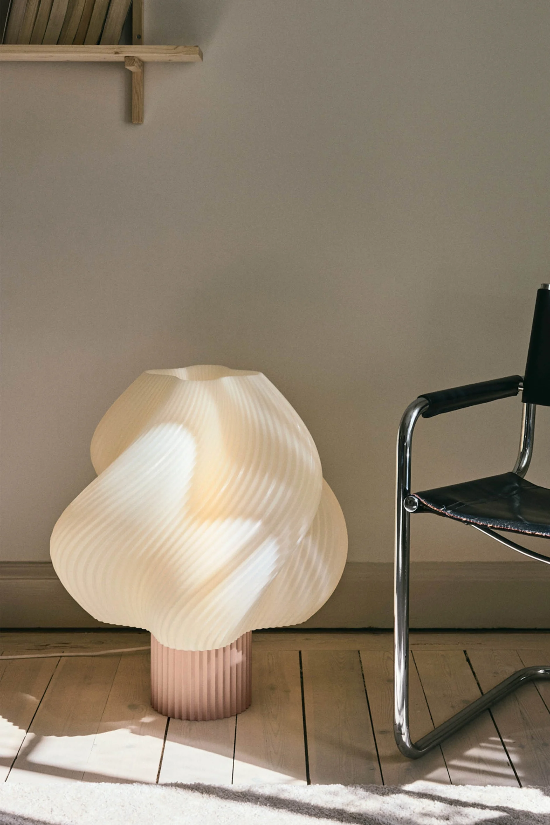 En skulptural, plisserad gräddfärgad golvlampa med en rosa bas står på ett trägolv bredvid en svart läder- och kromstol.