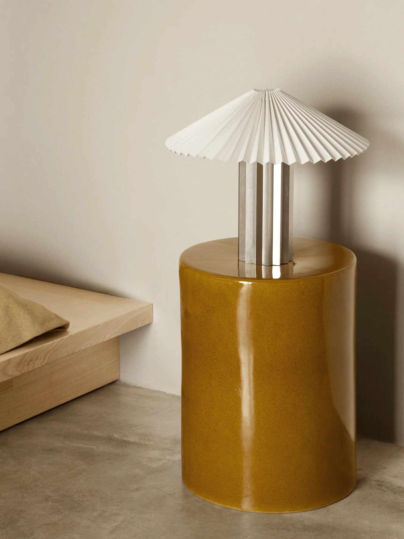 En bordslampa med vit plisserad skärm och silverfot står på ett glänsande senapsgult cylindriskt sidobord i ett minimalistiskt rum.