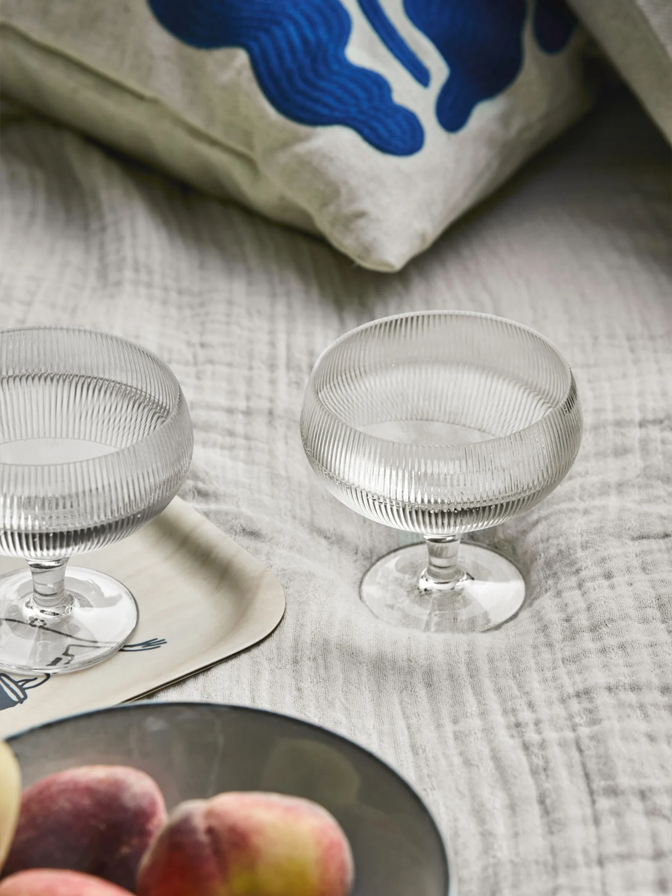 Två räfflade glasdessertskålar, en kudde med blå abstrakt konst, en träbricka och persikor på ett ljust tyg.