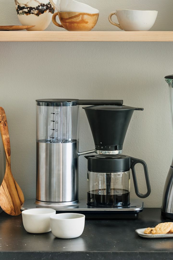 Du behöver också rätt apparater för din kaffebar hemma. Här ser du en klassisk filterkaffebryggare från Wilfa stå på en köksbänk, med en hylla med skålar och en brödrost ovanför.