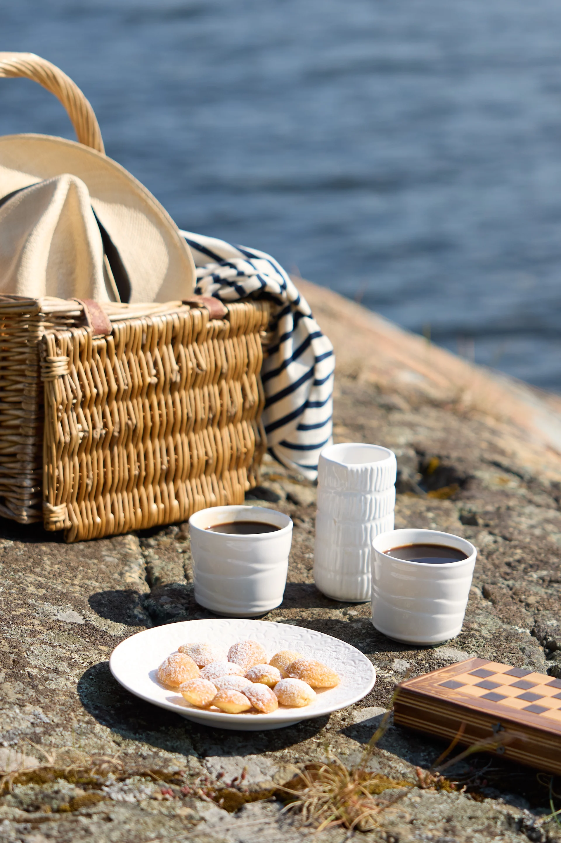 En picknick på klippig terräng vid vattnet, med kaffe, bakverk och ett schackspel.