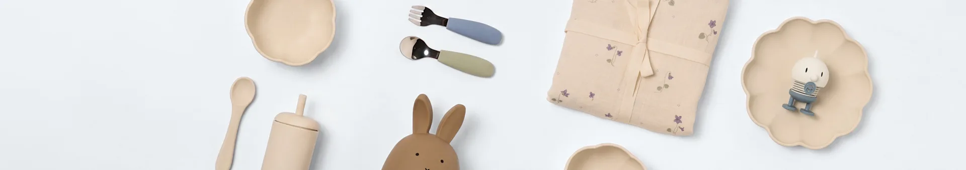 Babyartiklar på vitt: två krämfärgade skålar, blåa och gröna bestick, blommigt tyg, kaninleksak, träsked, beige mugg och liten figur på en vågig tallrik.